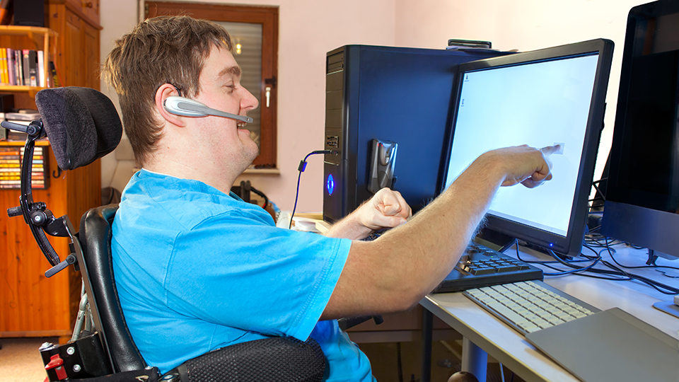 Person in a wheelchair uses a headset to control or interact with a computer, pointing at a screen in a home office setup.