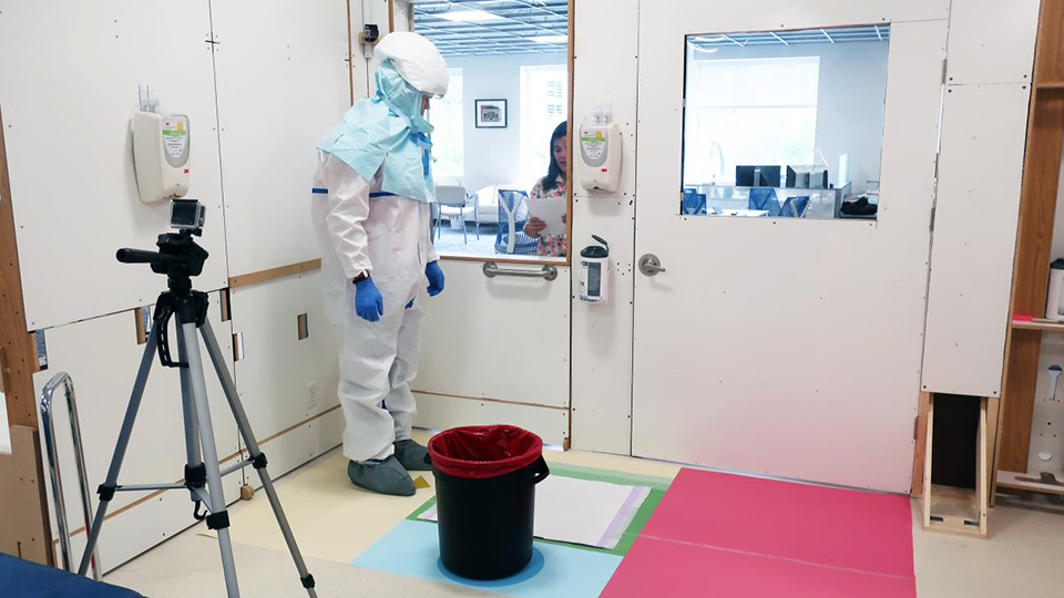 Person wearing full protective suit stands in a controlled lab room near a biohazard bin, with camera equipment and observation window visible.
