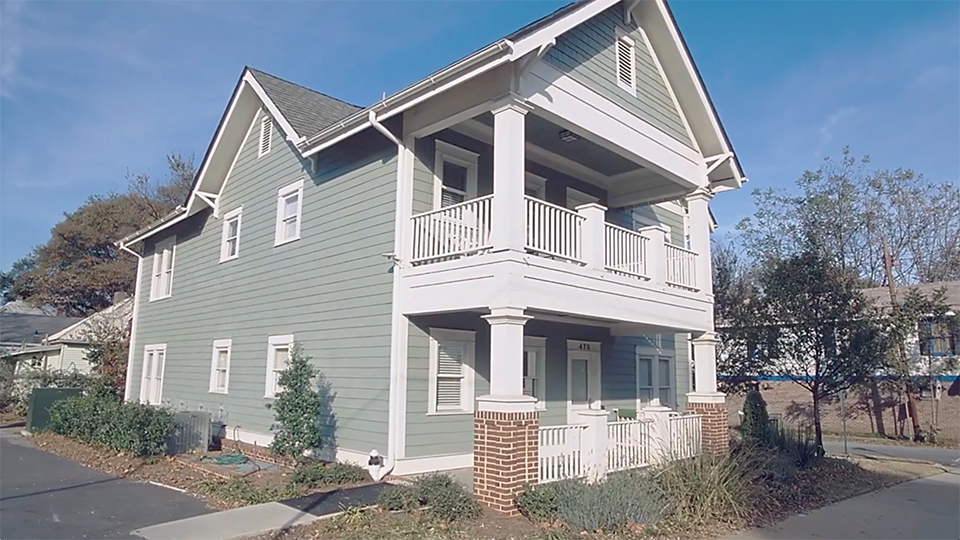 Two-story light green house with white trim, covered front porches on both levels, brick column bases, and landscaping along a sidewalk under a clear blue sky.