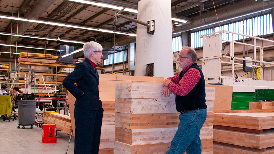 Two people stand across stacked wood panels, talking in a large workshop or fabrication space with industrial equipment.